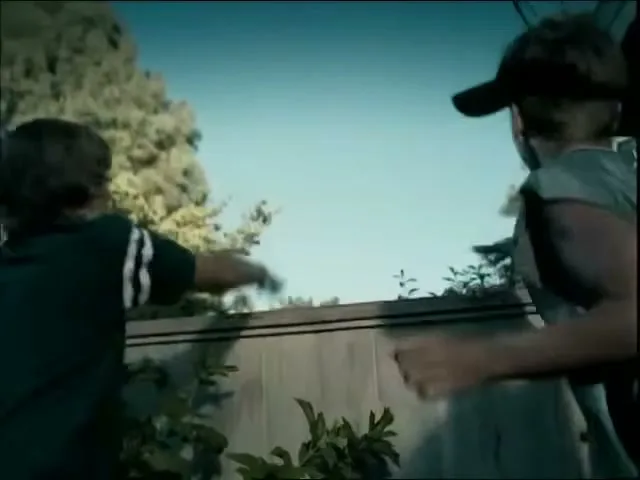 Three boys are playing in a backyard, with one boy being pulled over a fence by the other two, under a bright blue sky.