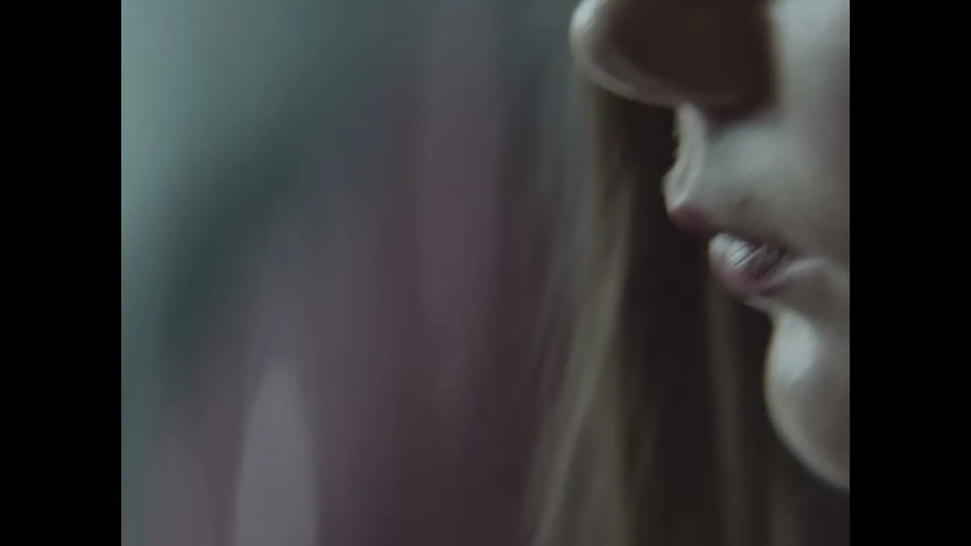 A young female with long brown hair looks down, her face partially obscured by shadow, in a close-up shot.