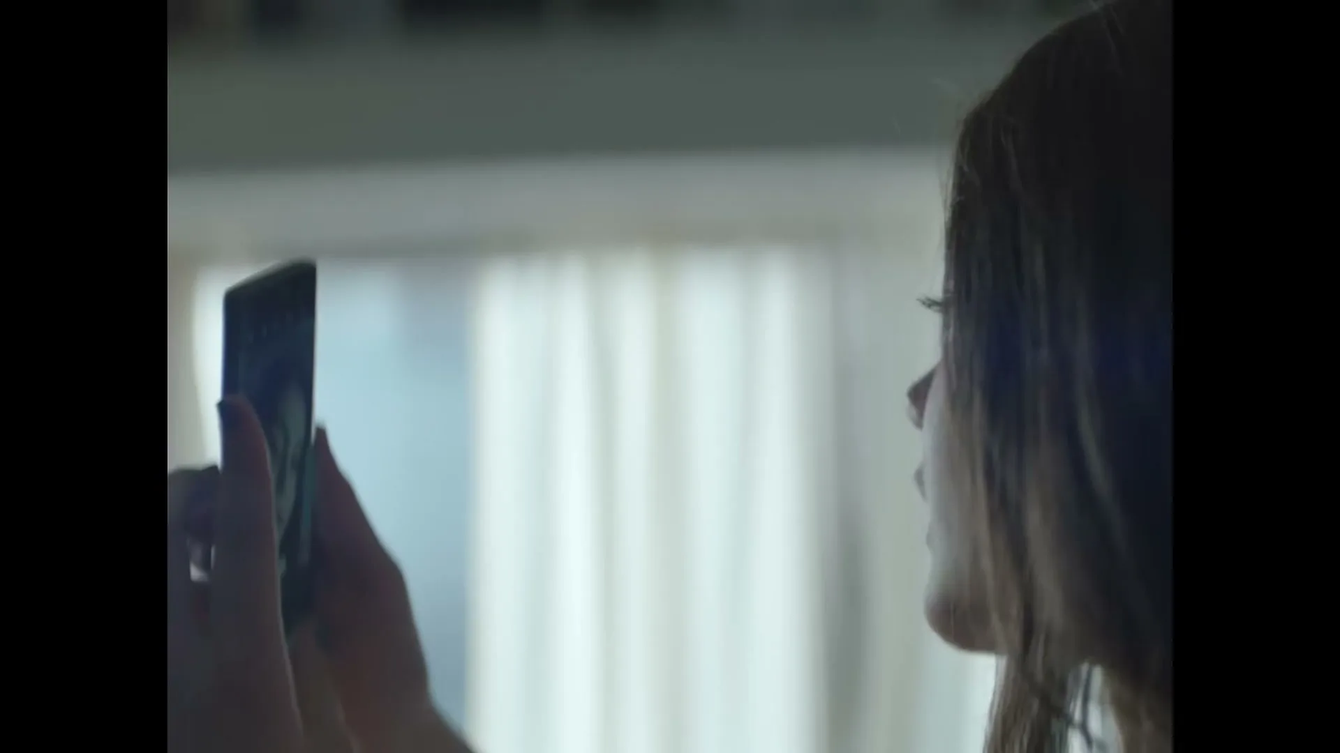 A young woman with long brown hair holds up a smartphone to take a selfie in a softly lit room.