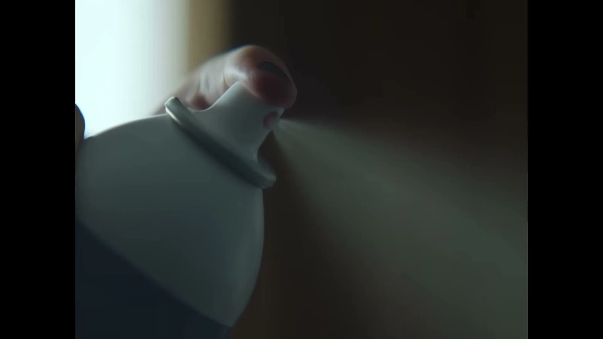 A hand with dark nail polish holds a white spray can, spraying a mist to the right, in a dimly lit interior.