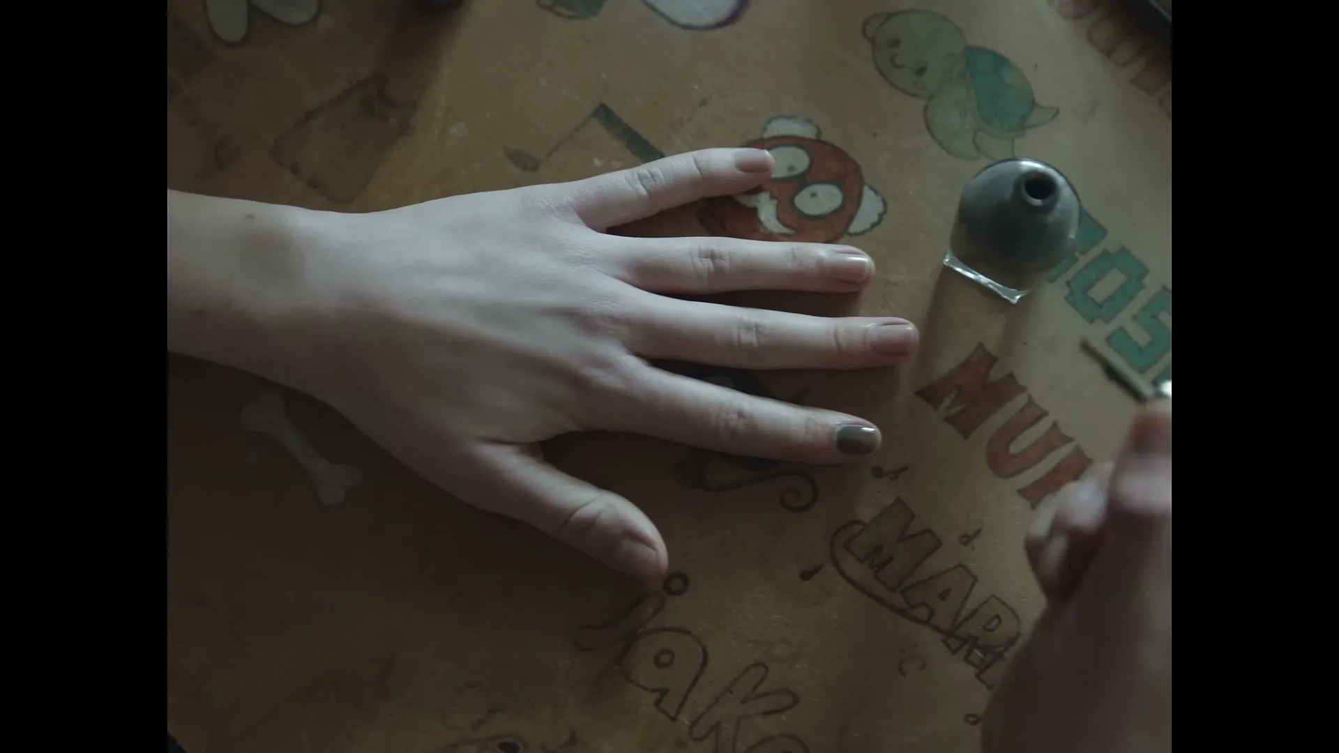 A young person's hand is seen getting their fingernails painted with brown nail polish on a patterned table.