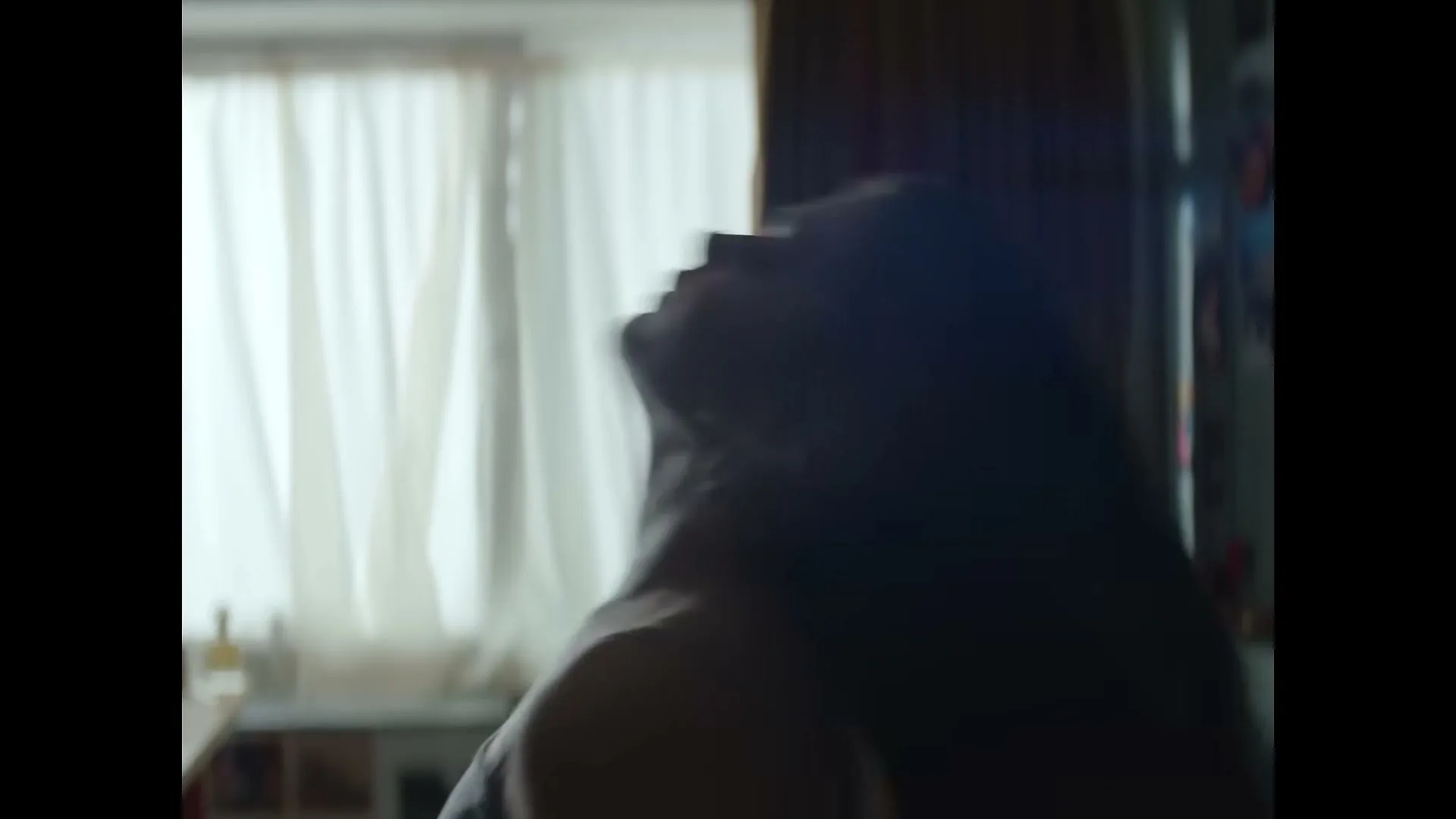 A young woman with long hair, in silhouette, pulls her hair into a ponytail in her bedroom during the day.