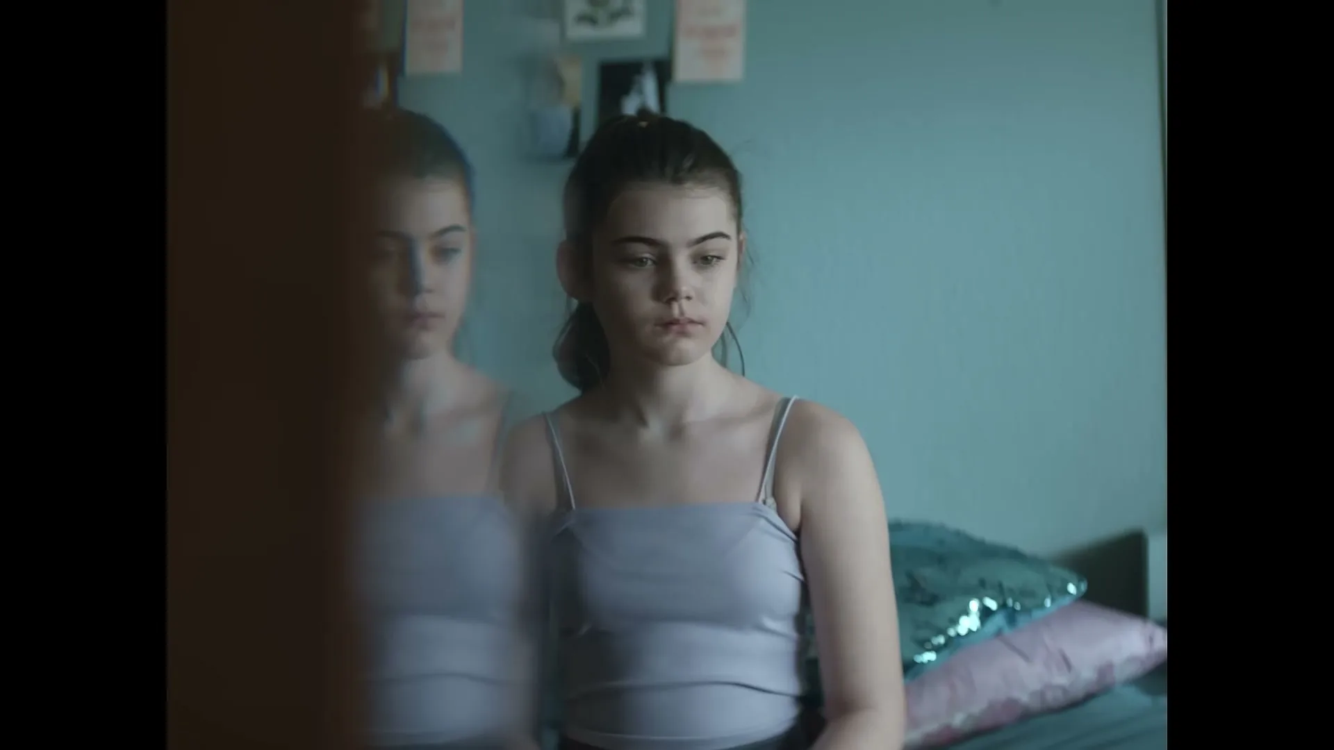 A young girl with dark hair in a light purple top looks at her reflection in a mirror in her bedroom, appearing sad.