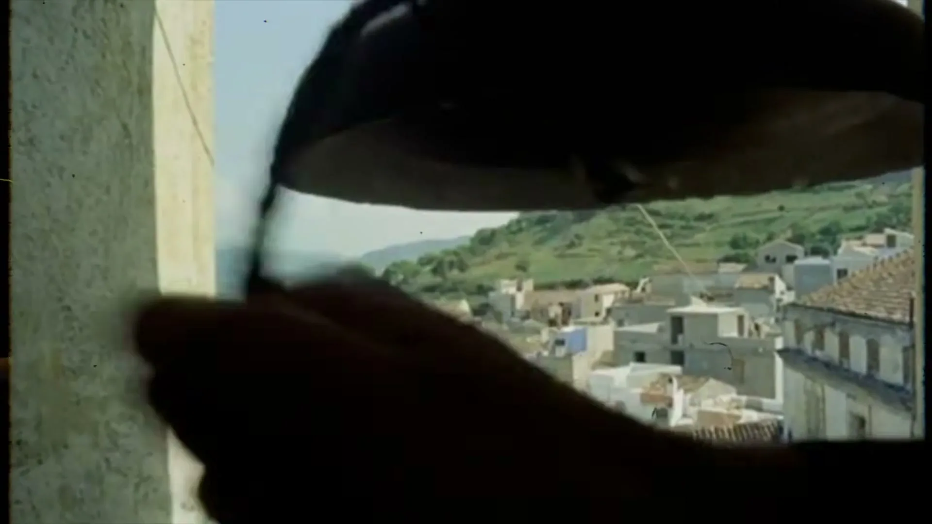 A silhouetted hand rings a large bell in a bell tower, overlooking a village nestled in a green, hilly landscape under a bright sky.