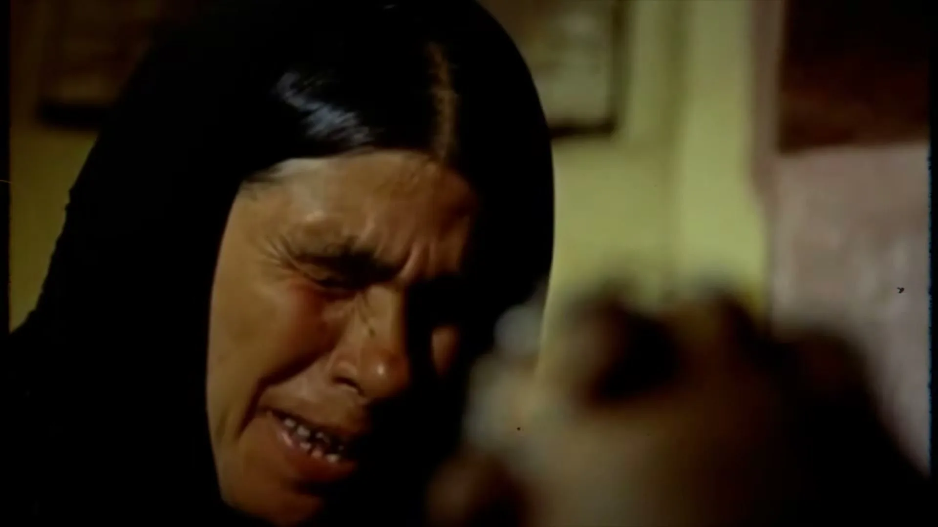 A distraught older woman in a black headscarf cries intensely, her face contorted in grief, as she holds a string of beads.