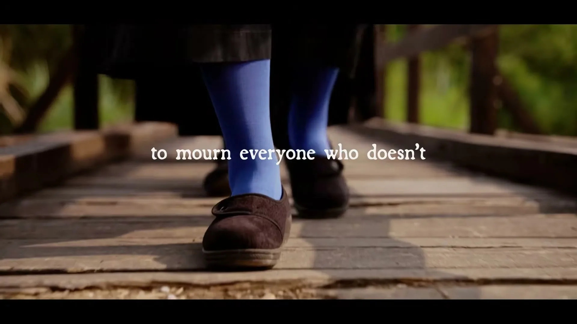 A person wearing blue tights and brown shoes walks on a wooden bridge, with text overlayed on the screen.