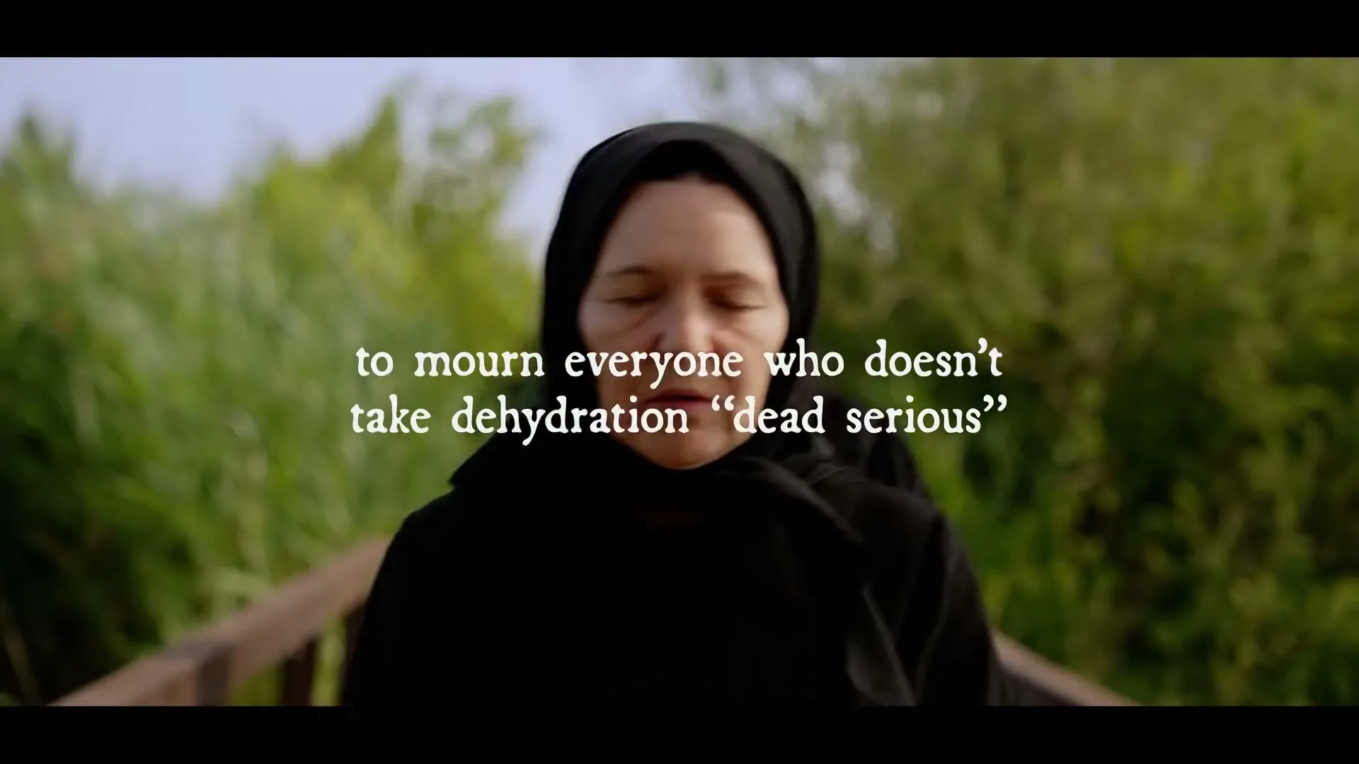 Two women in black hijabs walk slowly on a wooden bridge, with green foliage in the background, as text appears on screen.