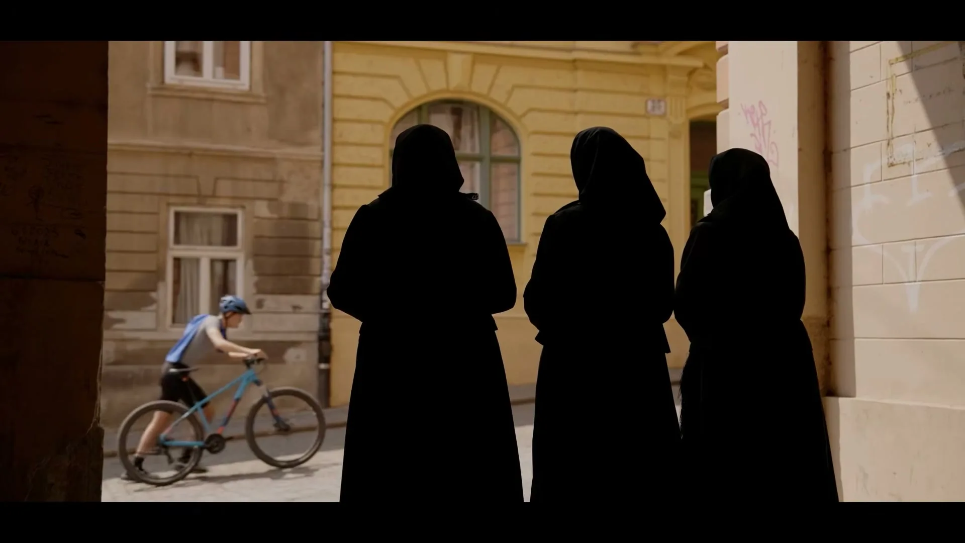 Three women in black robes stand in silhouette in an alleyway, watching a boy on a bicycle ride past on a sunny day.