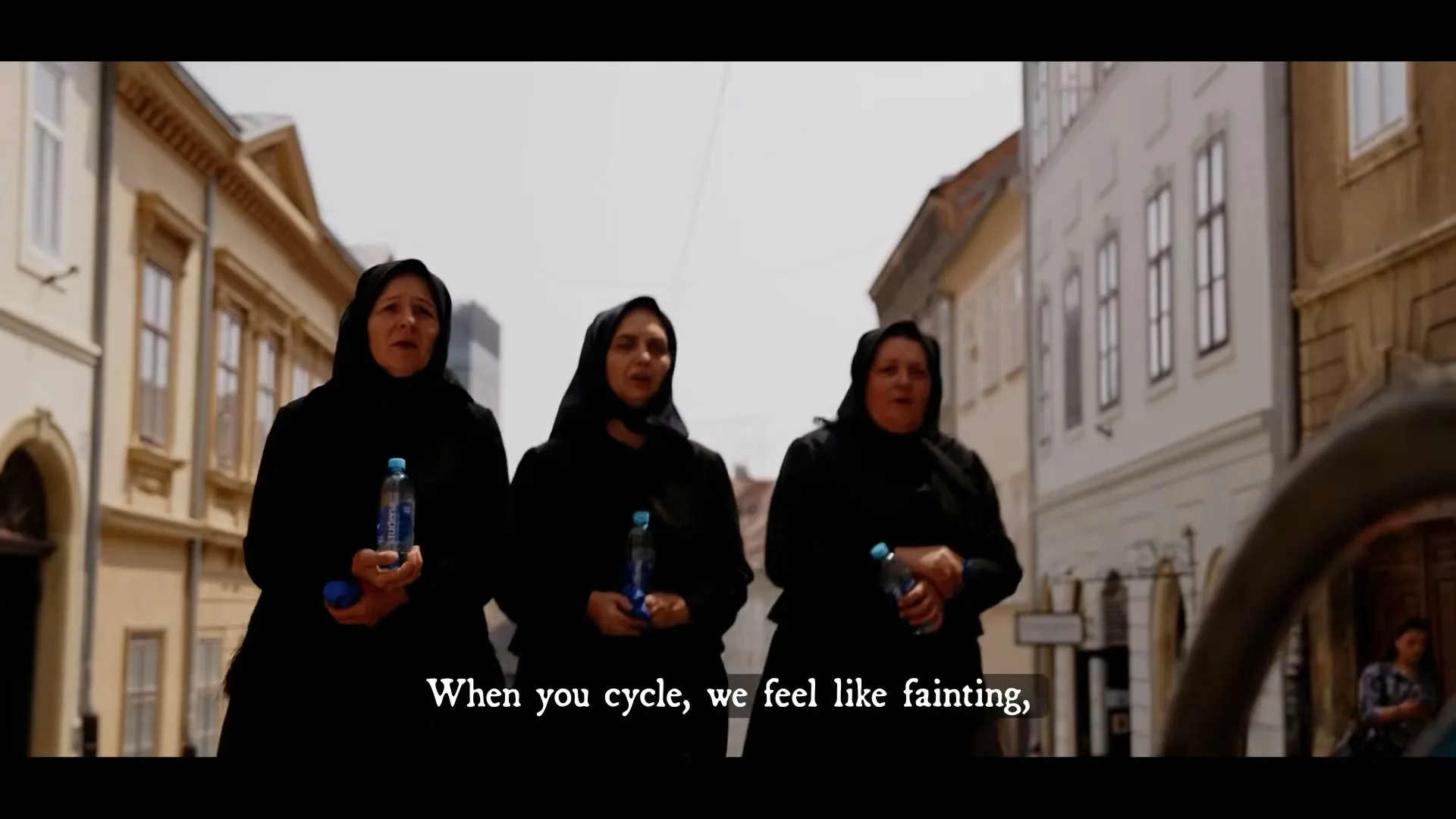 A young male cyclist, wearing a helmet and a blue cape, looks back at three older women in black dresses and headscarves, holding water bottles on a sunny street.