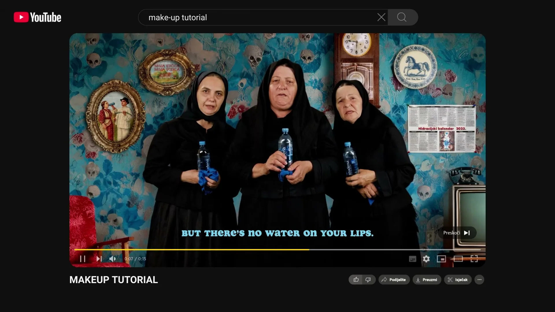 Three older women in black dresses and headscarves hold water bottles, standing against a blue floral wallpaper with framed pictures and a clock.