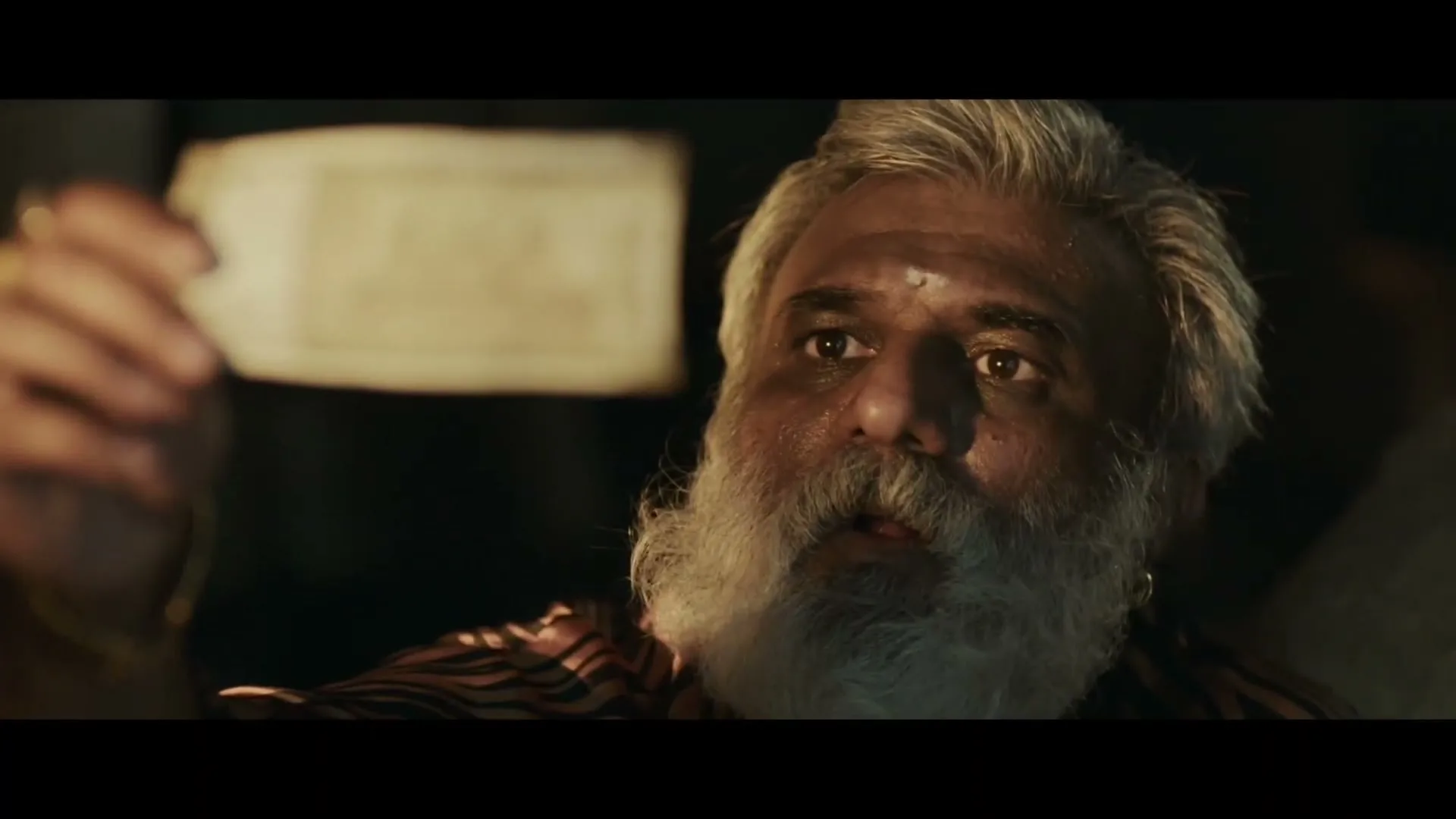 A man with a white beard and dark skin holds up a 500 Rupee note, looking surprised in a dimly lit interior.