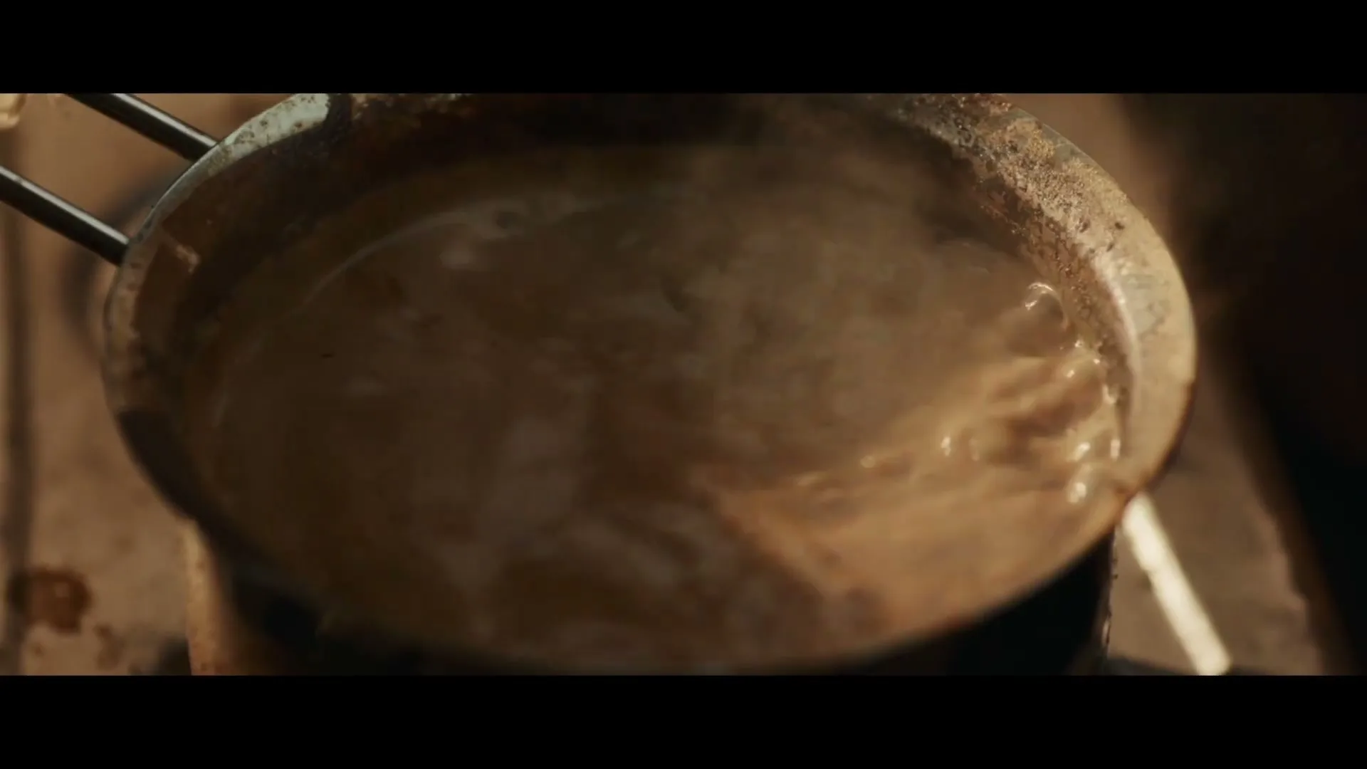 A close-up shot shows a metal pot filled with a light brown liquid boiling on a stove, with steam rising from the surface.