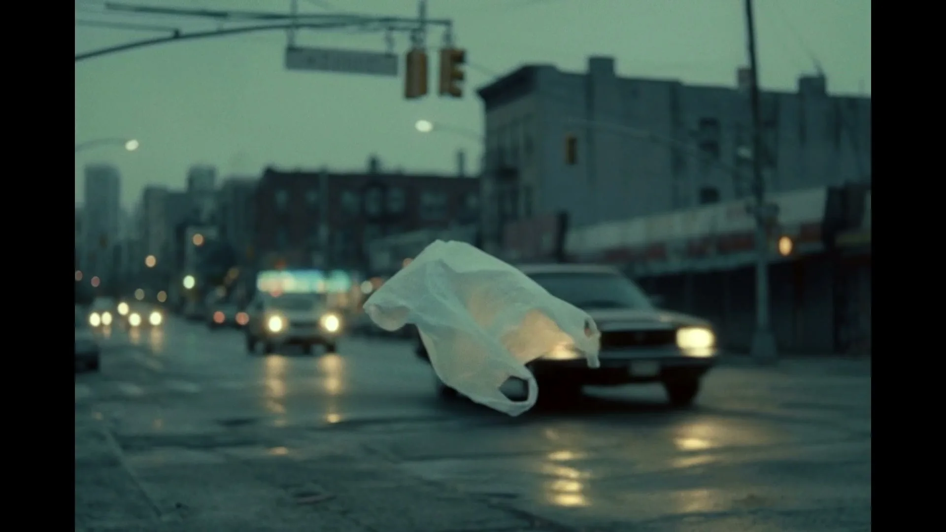 A white plastic bag floats in the air over a wet city street with cars driving by and buildings in the background on a cloudy day.