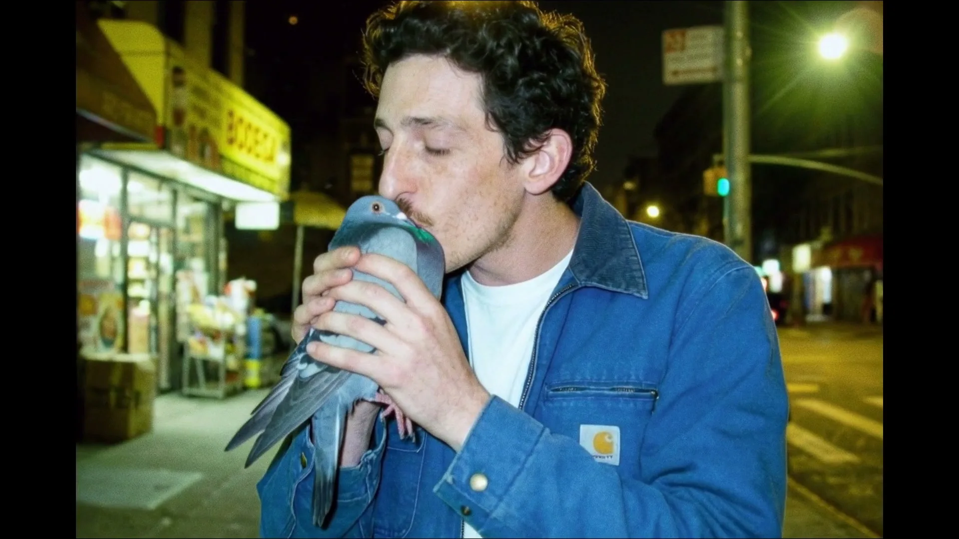A man with a mustache, wearing a blue jacket, holds a pigeon to his face and kisses it on a city street at night.
