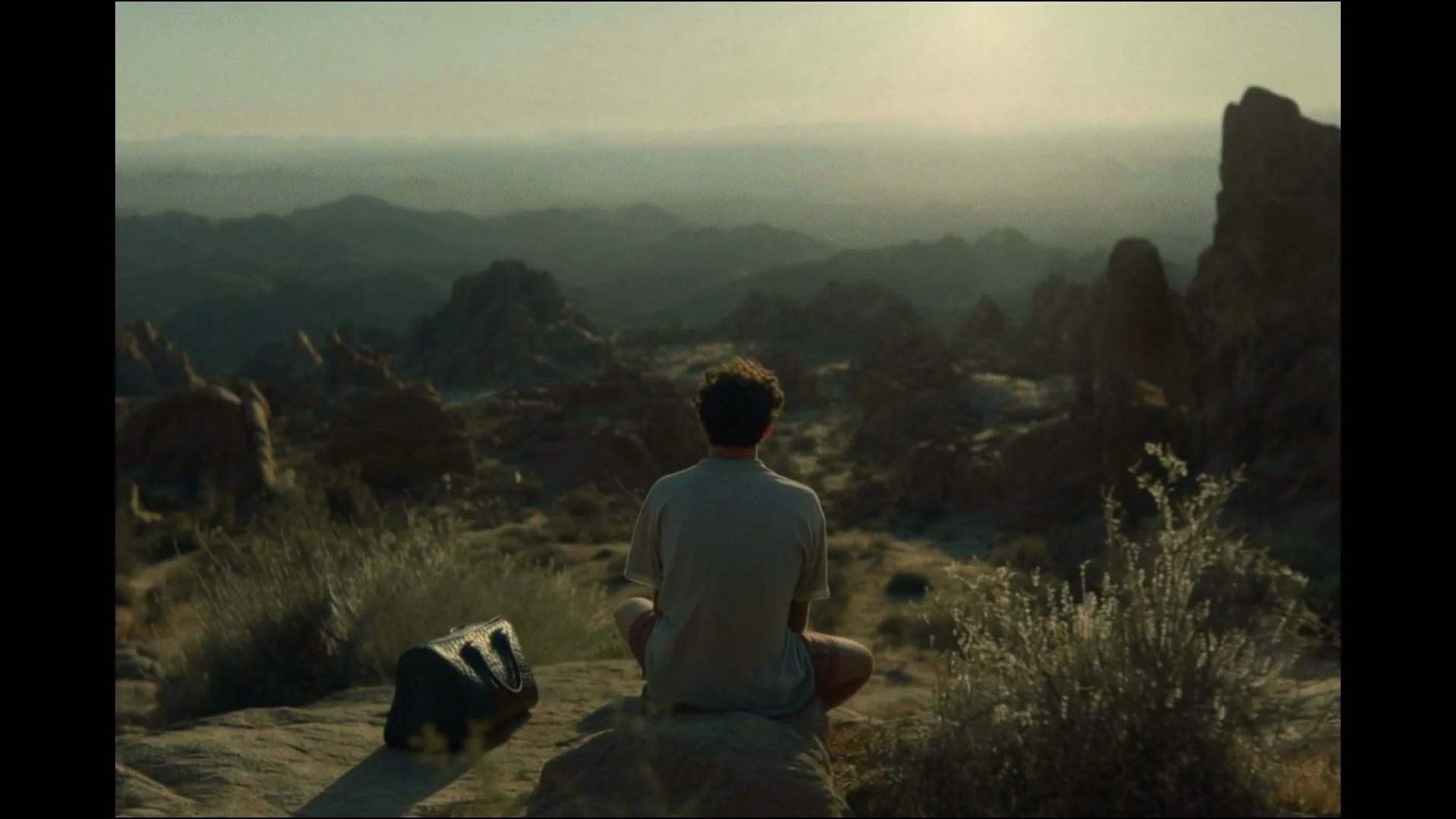 A man with curly hair sits cross-legged on a rock, looking out at a vast desert landscape with mountains under a hazy sky.