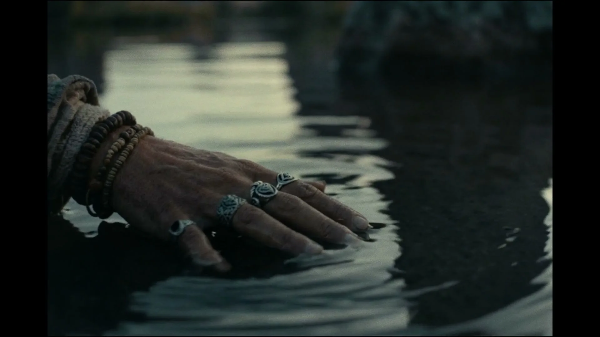 A person's hand, adorned with rings and bracelets, gently touches the surface of calm water, creating small ripples.