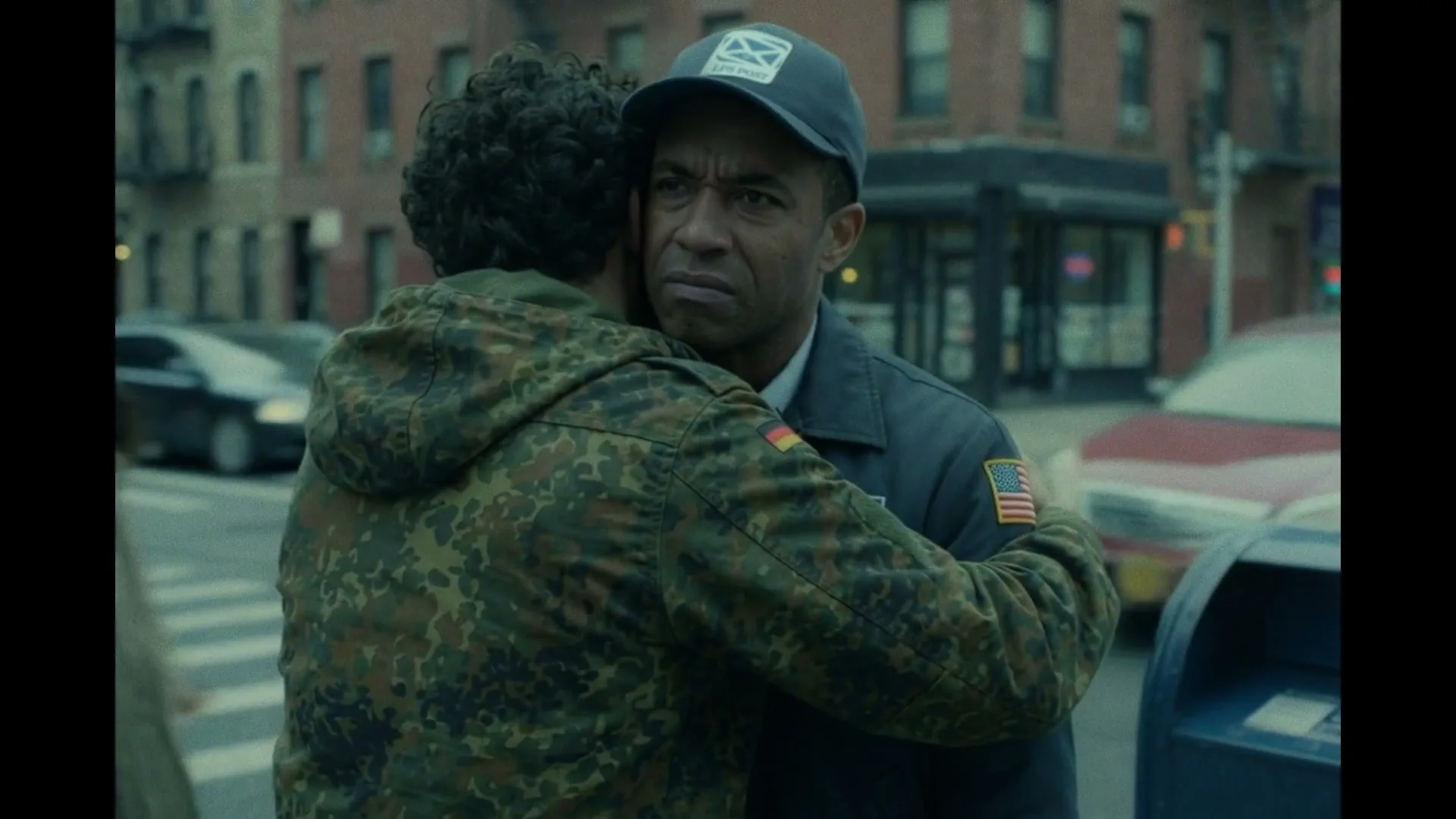 A man in a camouflage jacket hugs a mailman in uniform on a city street during the day.