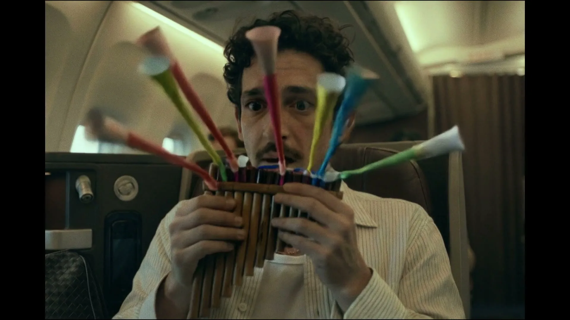 A man with curly hair and a mustache plays a pan flute on an airplane, then party blowers pop out of it, surprising him.
