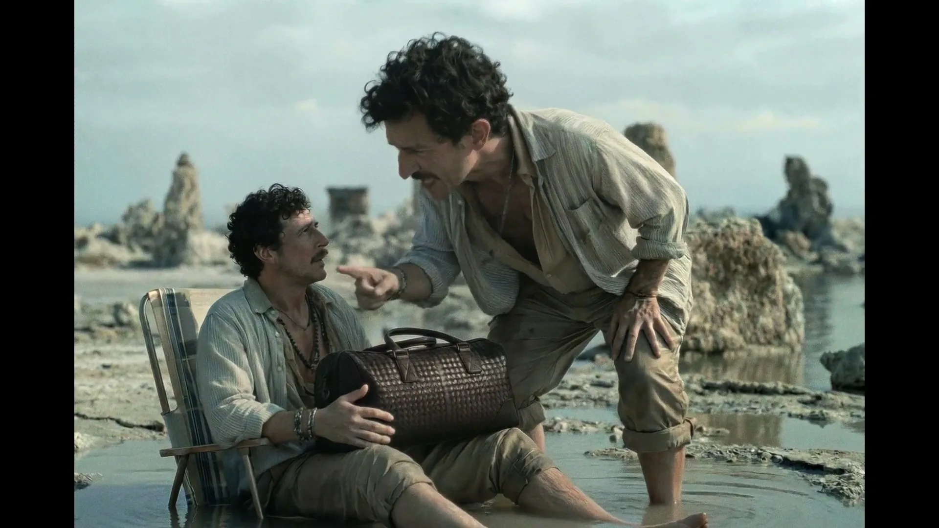 Two men, one sitting in a chair in shallow water holding a duffel bag, the other standing and pointing at him, in a desolate, rocky landscape.