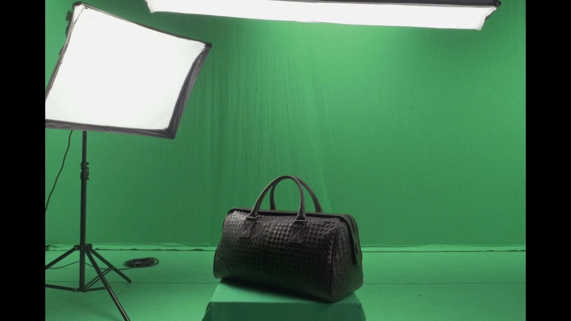 A black woven leather duffel bag sits on a green pedestal in a studio with a green screen background and studio lights.