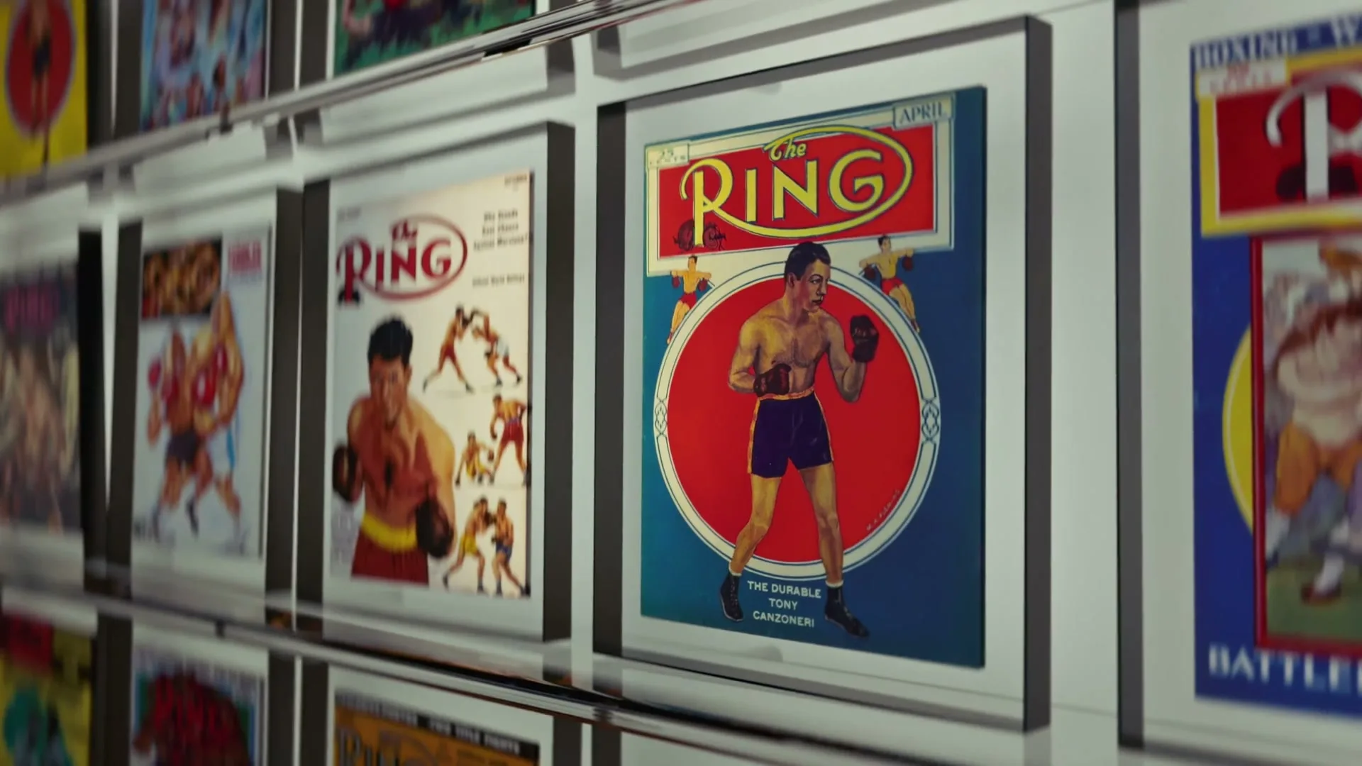 A camera moves along a wall displaying framed vintage boxing magazine covers, with "The Ring" magazine prominently featured.