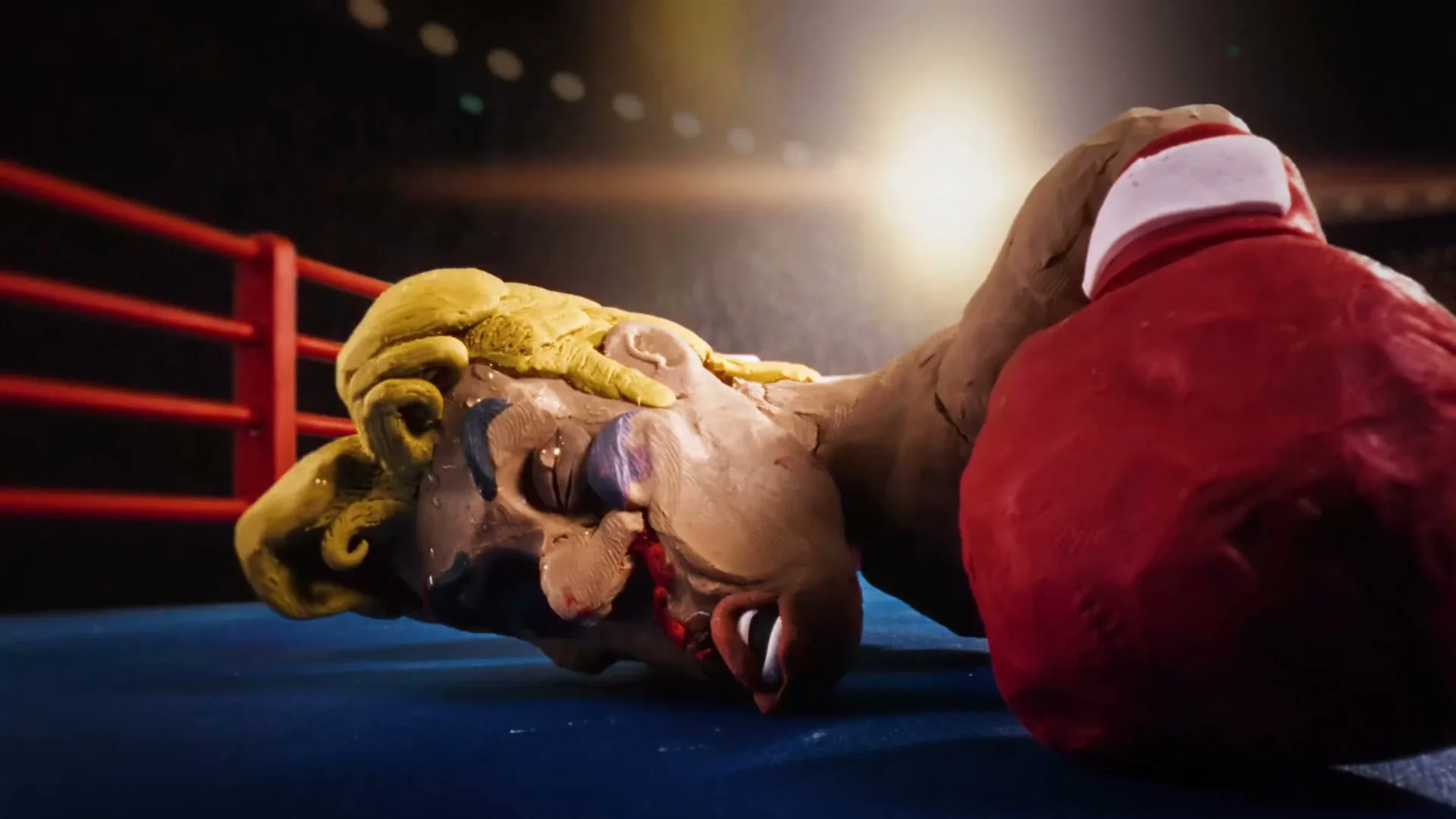 A claymation boxer with blonde hair and a red glove is knocked out on the blue mat of a boxing ring, with red and white ropes.