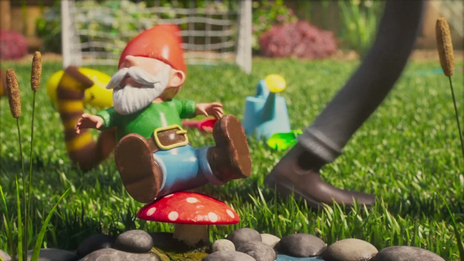A garden gnome with a red hat and white beard sits on a mushroom as a person's legs walk past in a sunny backyard.