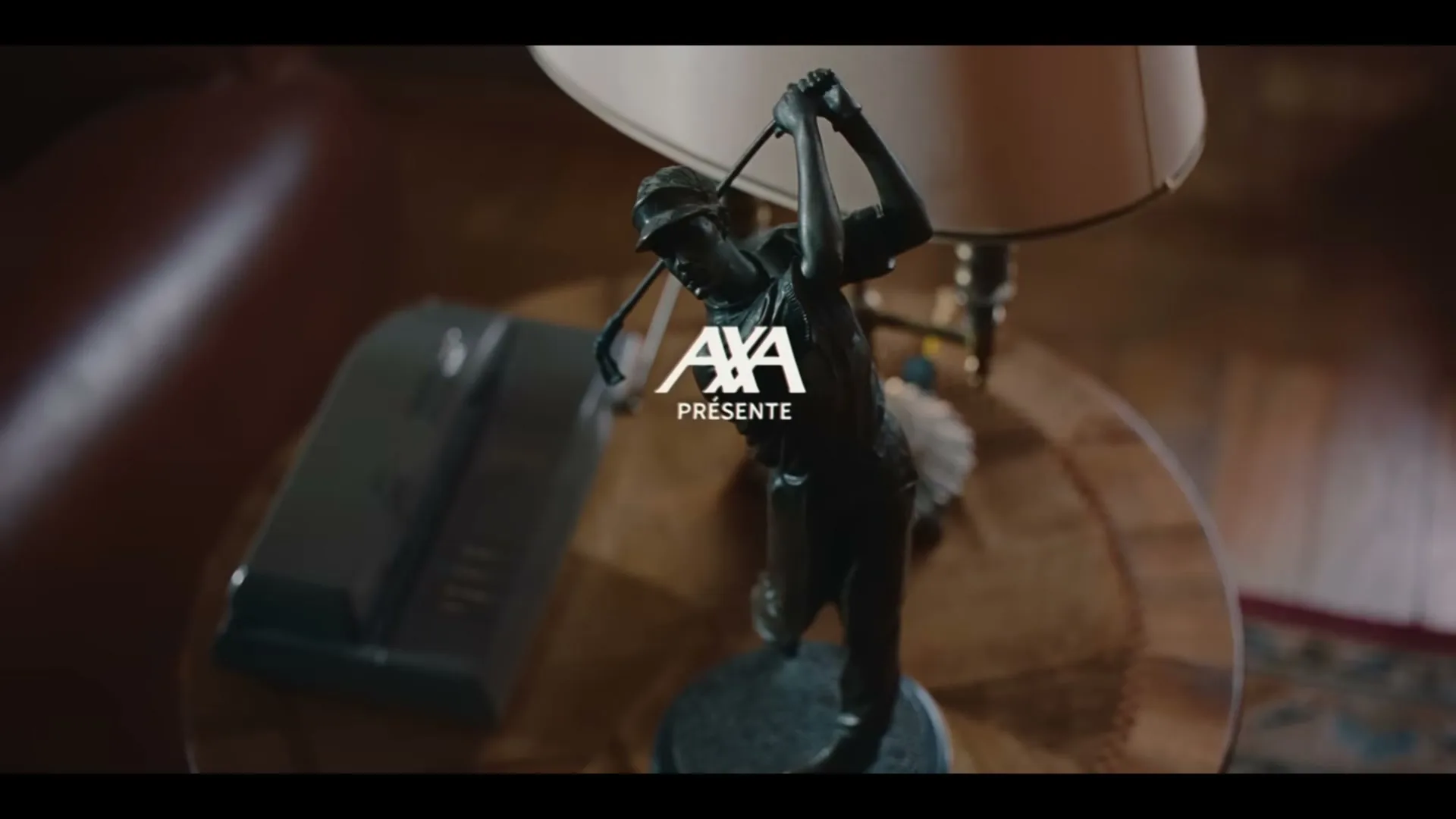A bronze statue of a male golfer mid-swing stands on a polished wooden table next to a lamp, with the AXA logo superimposed.
