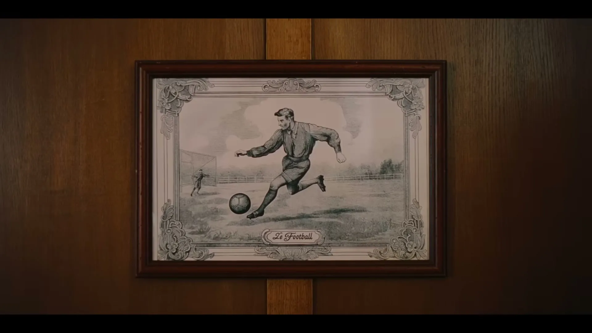 A framed, vintage-style illustration of a male soccer player kicking a ball on a field hangs on a wooden wall.
