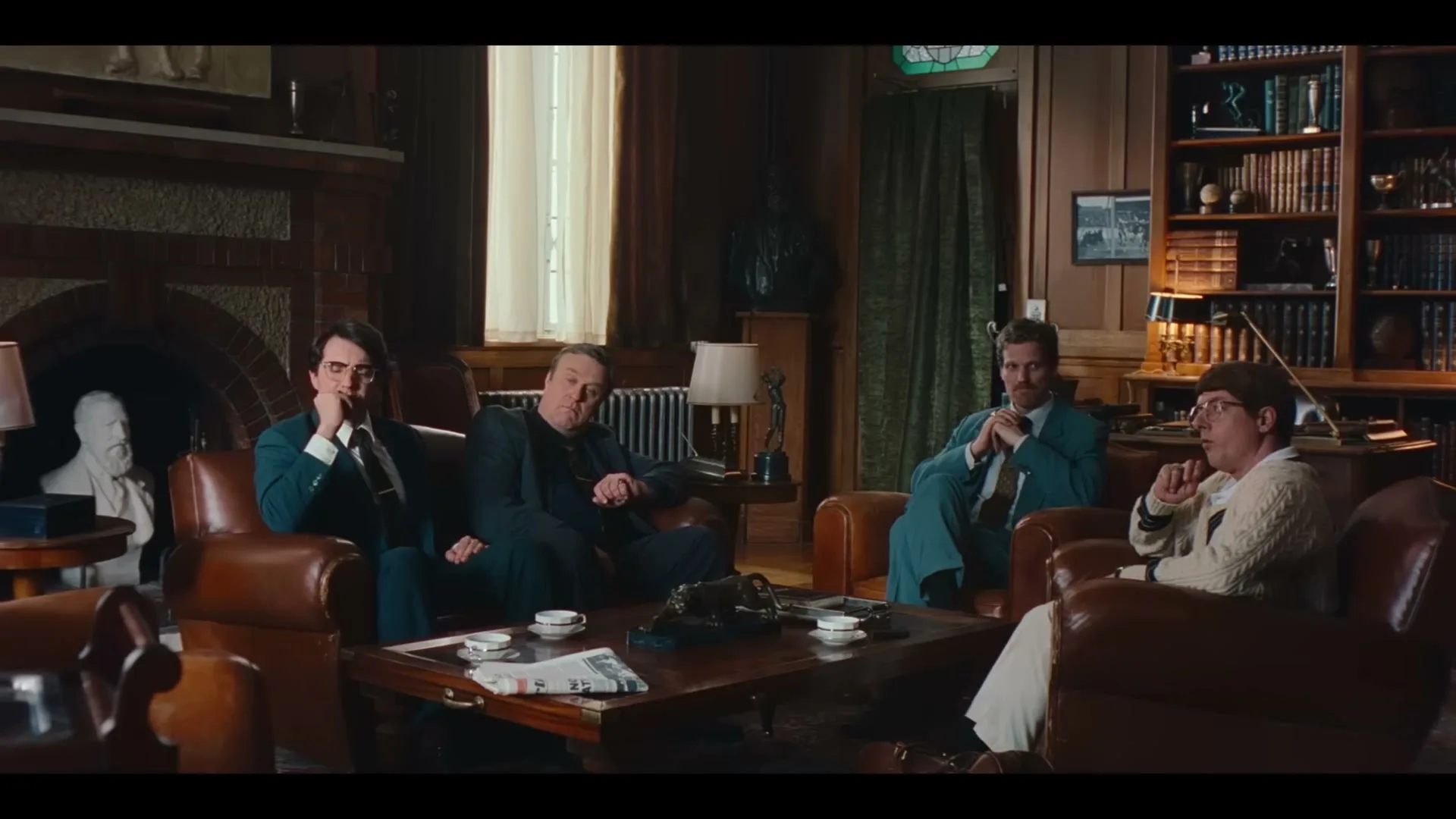 Four men in suits sit in leather armchairs around a coffee table in a wood-paneled study, listening intently.