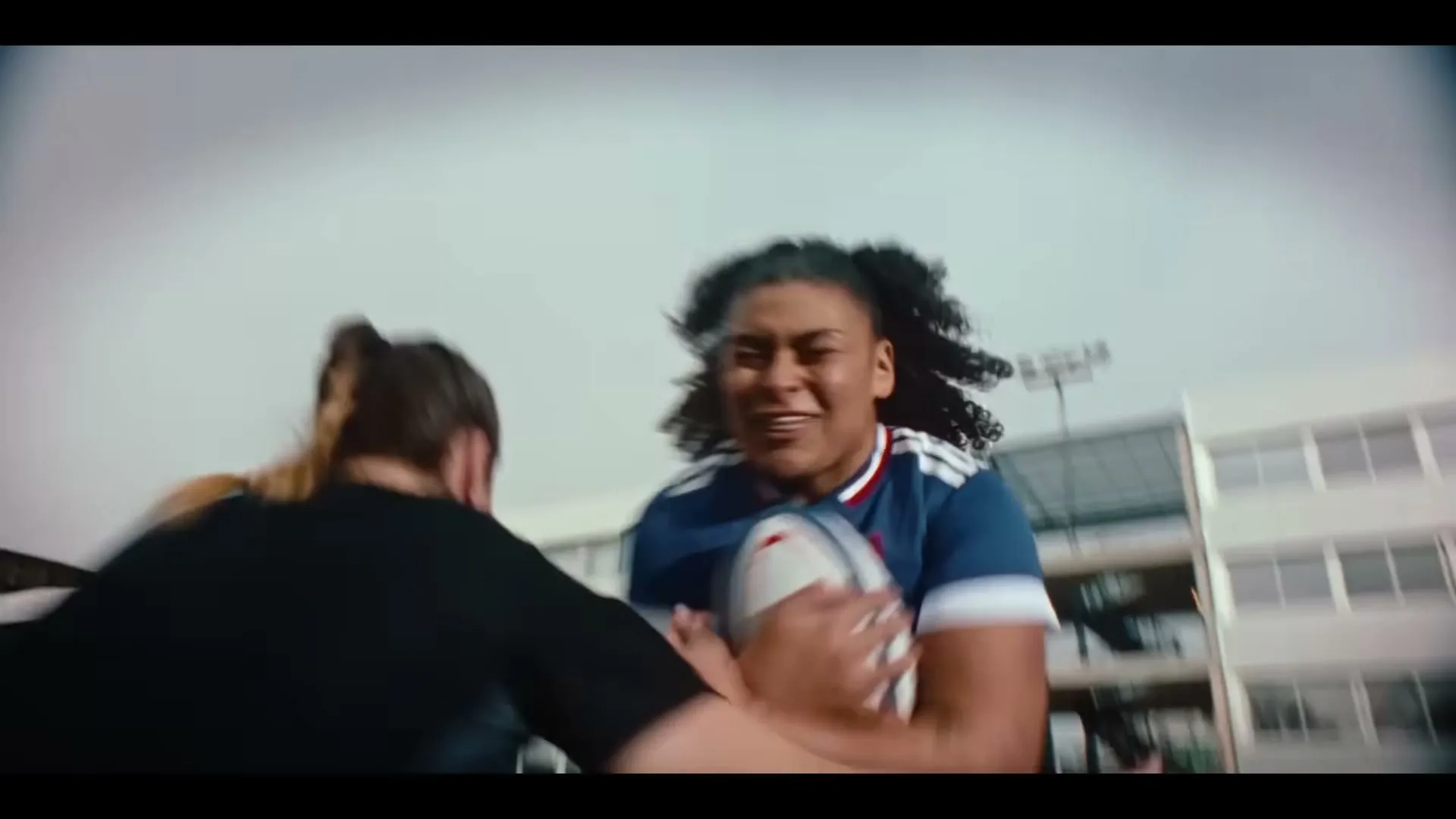 A female rugby player in a blue jersey runs with the ball, being tackled by an opponent on a sports field during the day.