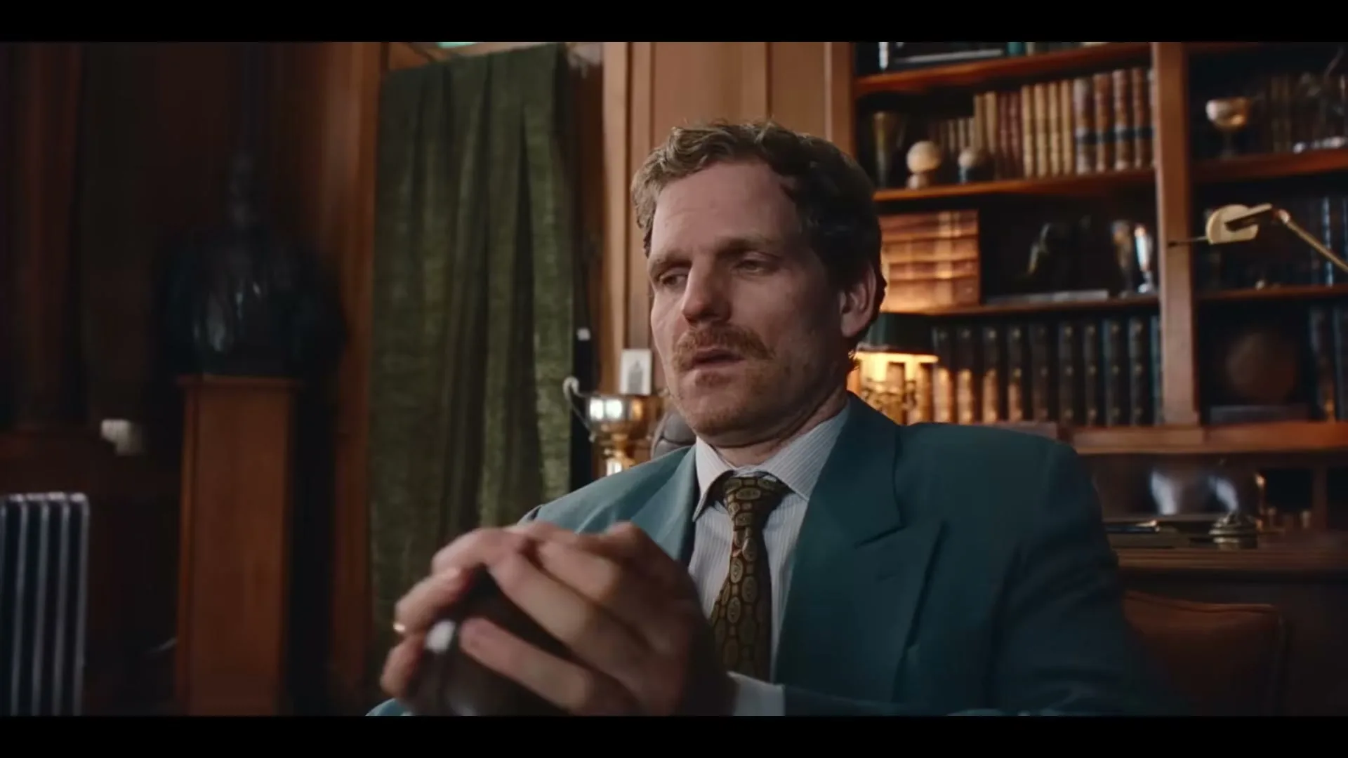 A man in his 30s, with a mustache and wearing a suit, sits at a desk in a wood-paneled office, looking surprised and then thoughtful.