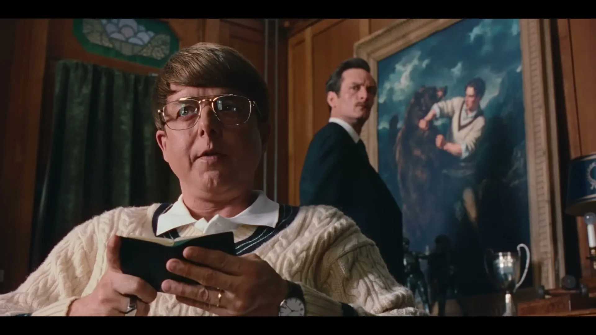 A middle-aged man with a bowl cut and glasses, wearing a cream cable-knit sweater, sits at a desk, holding a small black book and looking surprised, while another man stands behind him in a wood-paneled room.