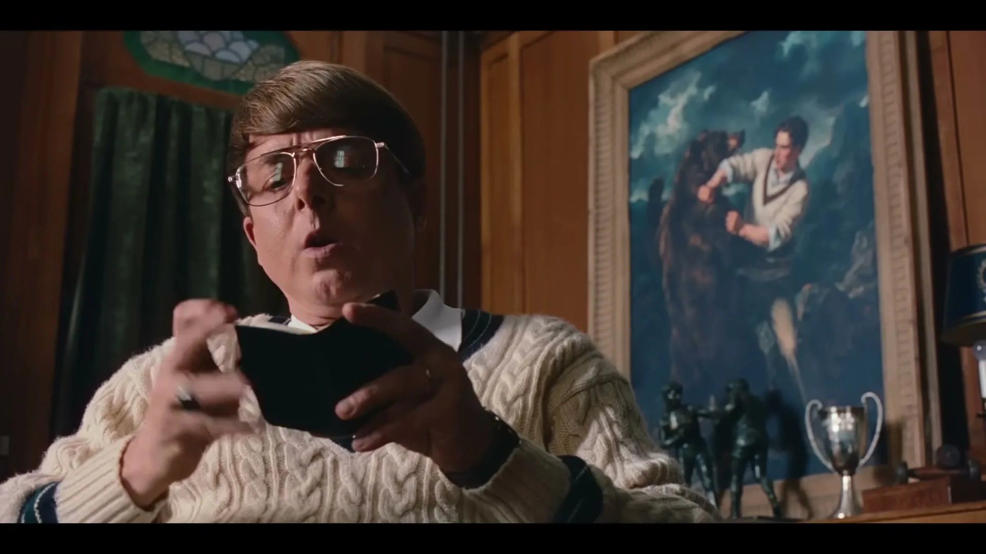 A man with a bowl cut and glasses, wearing a cable-knit sweater, holds a small book and speaks in a wood-paneled office.