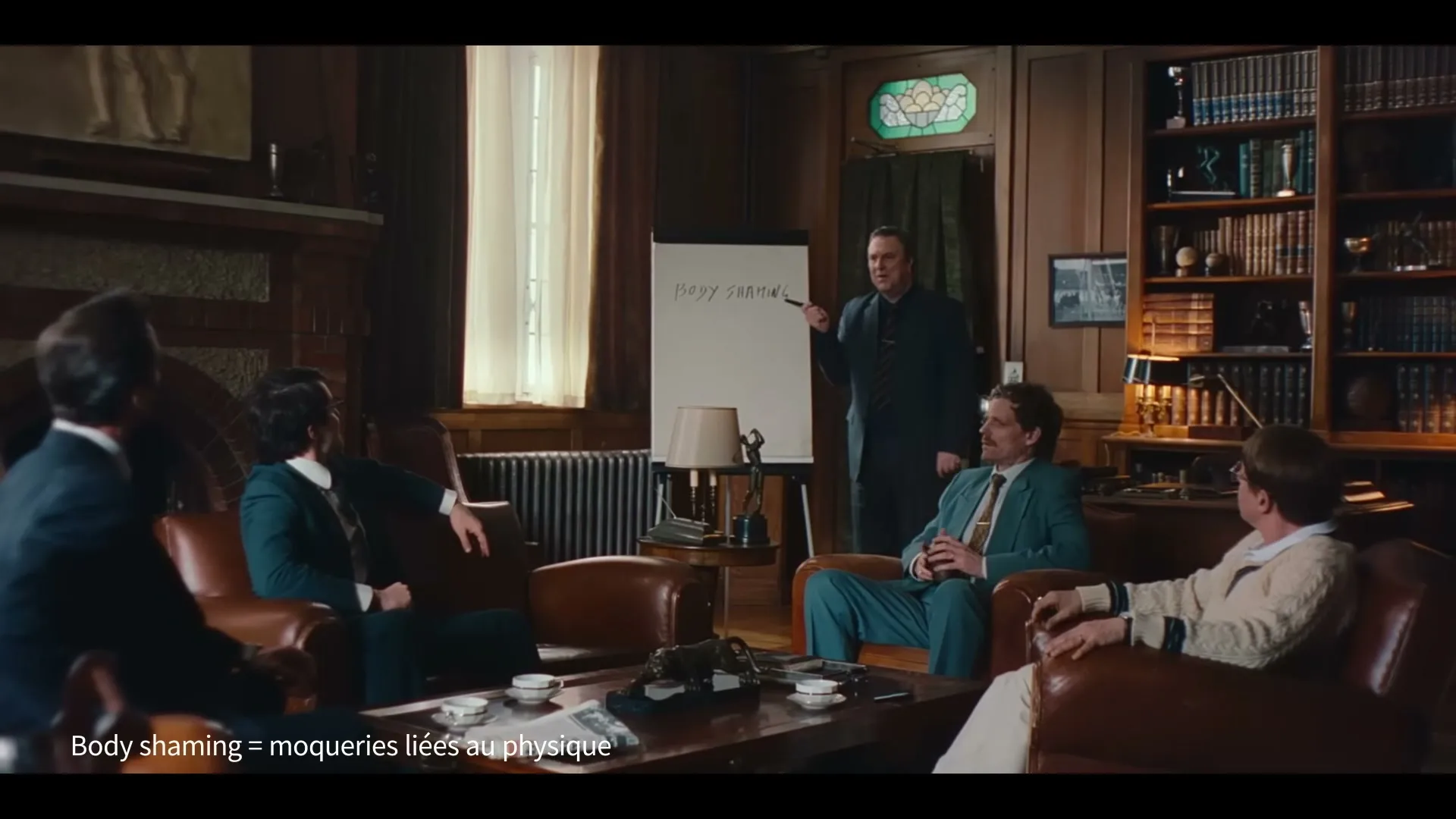 A group of men in suits sit in a wood-paneled room, listening to another man point at a whiteboard that says "BODY SHAMING."
