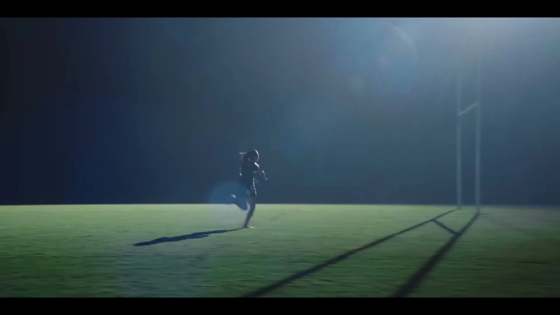 A female rugby player runs across a dark, floodlit rugby field at night, with goalposts visible on the right.