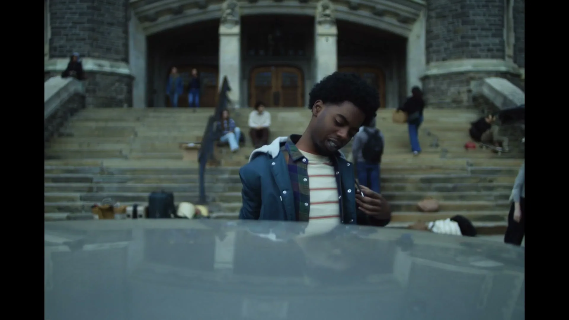 A young Black man with an afro leans over the roof of a car, adjusting his backpack, with a stone building and people on steps in the background.