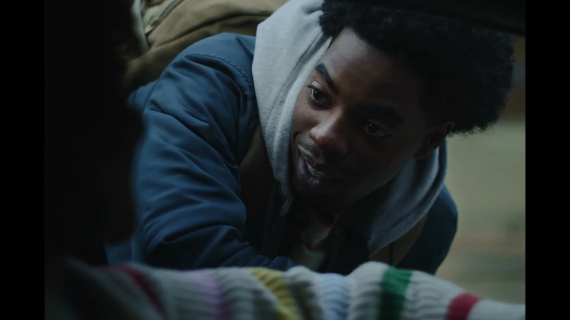 A young Black man in a blue jacket and grey hoodie leans into a car, smiling and talking to someone off-screen.
