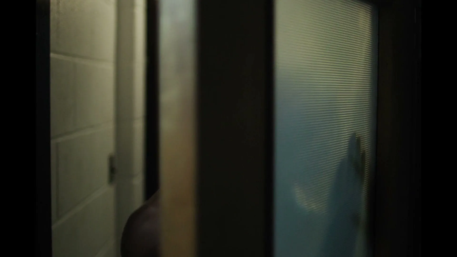 A young man with an afro and a silver chain opens a bathroom door, looking over his shoulder with a worried expression in a dimly lit hallway.