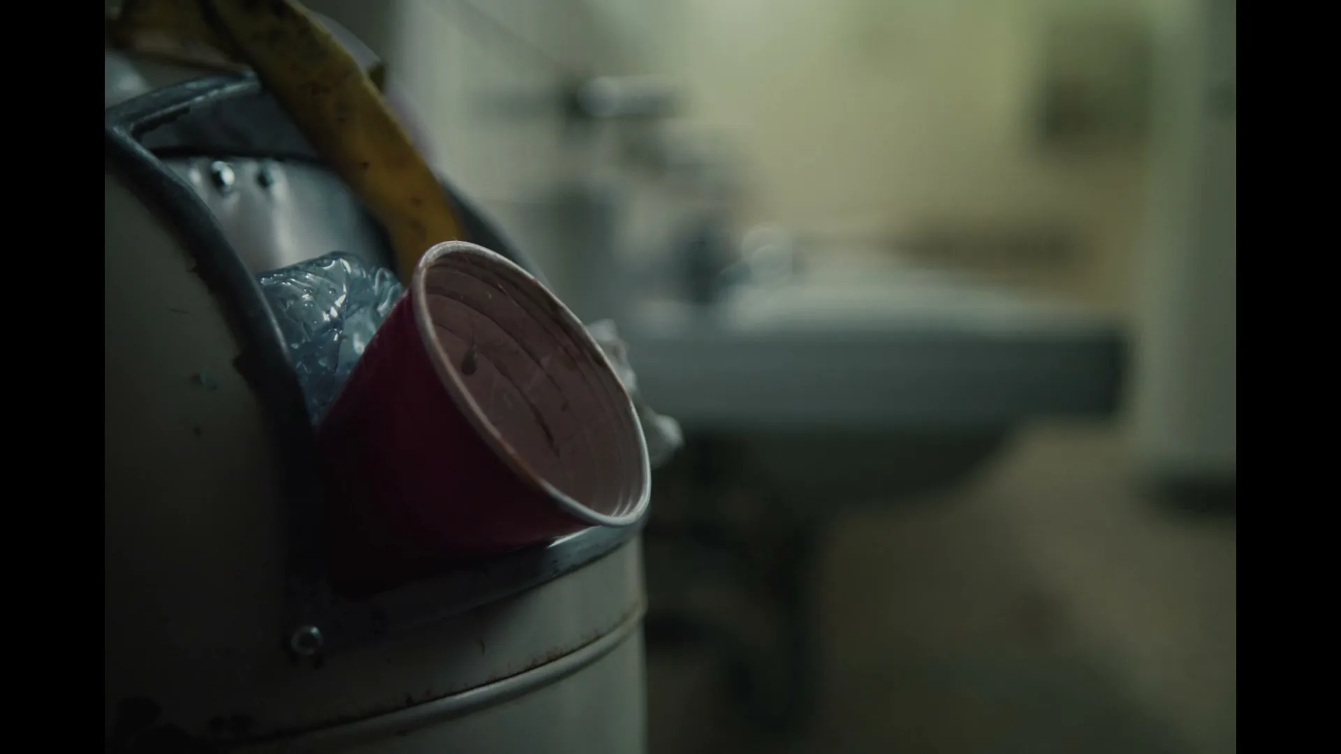 A close-up shot of a red plastic cup overflowing from a trash can, with a blurry bathroom sink in the background, creating a sense of neglect.