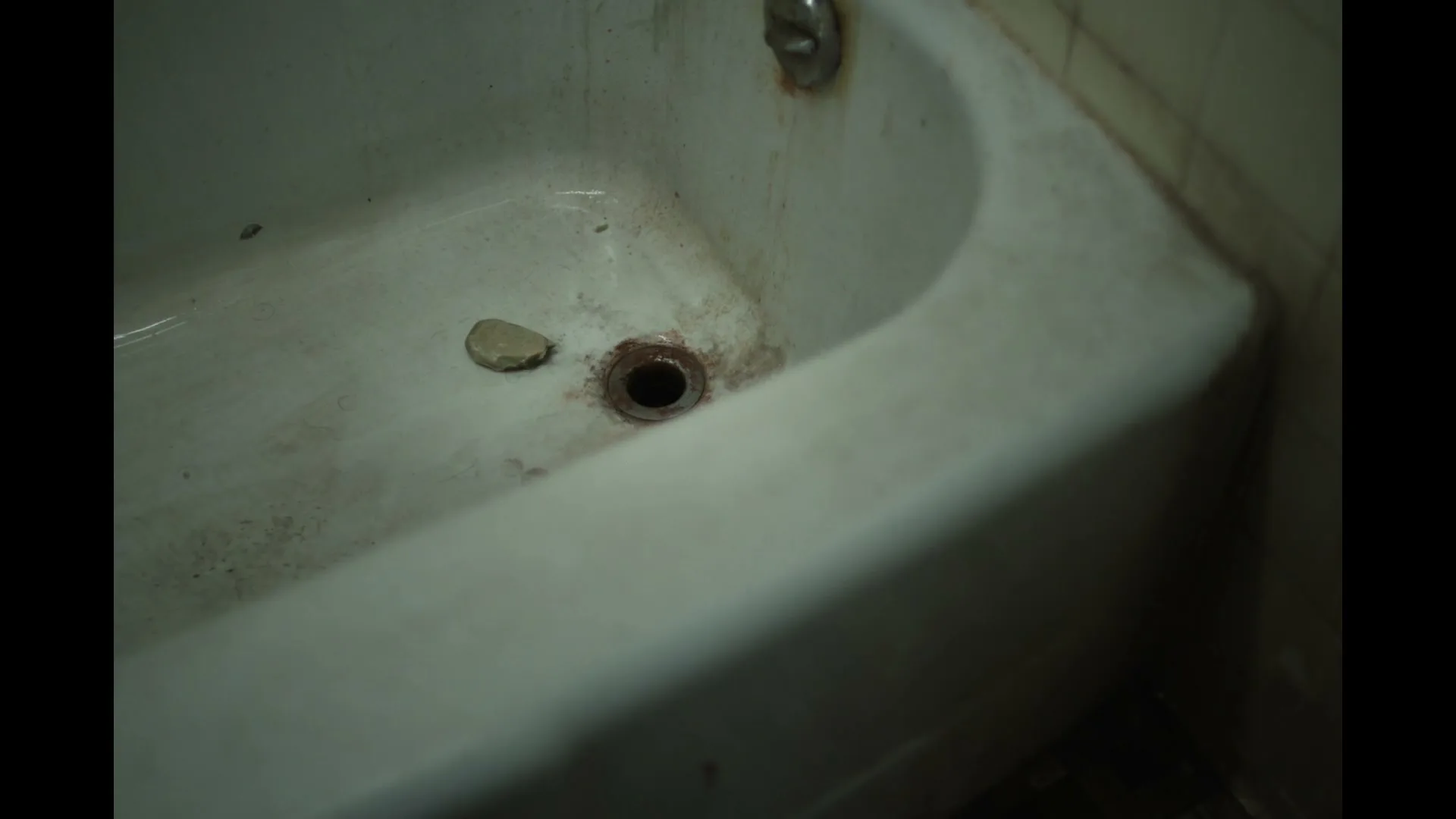 A dirty, old bathtub with a dark drain and a small, light-colored stone resting near it, in a dimly lit bathroom.