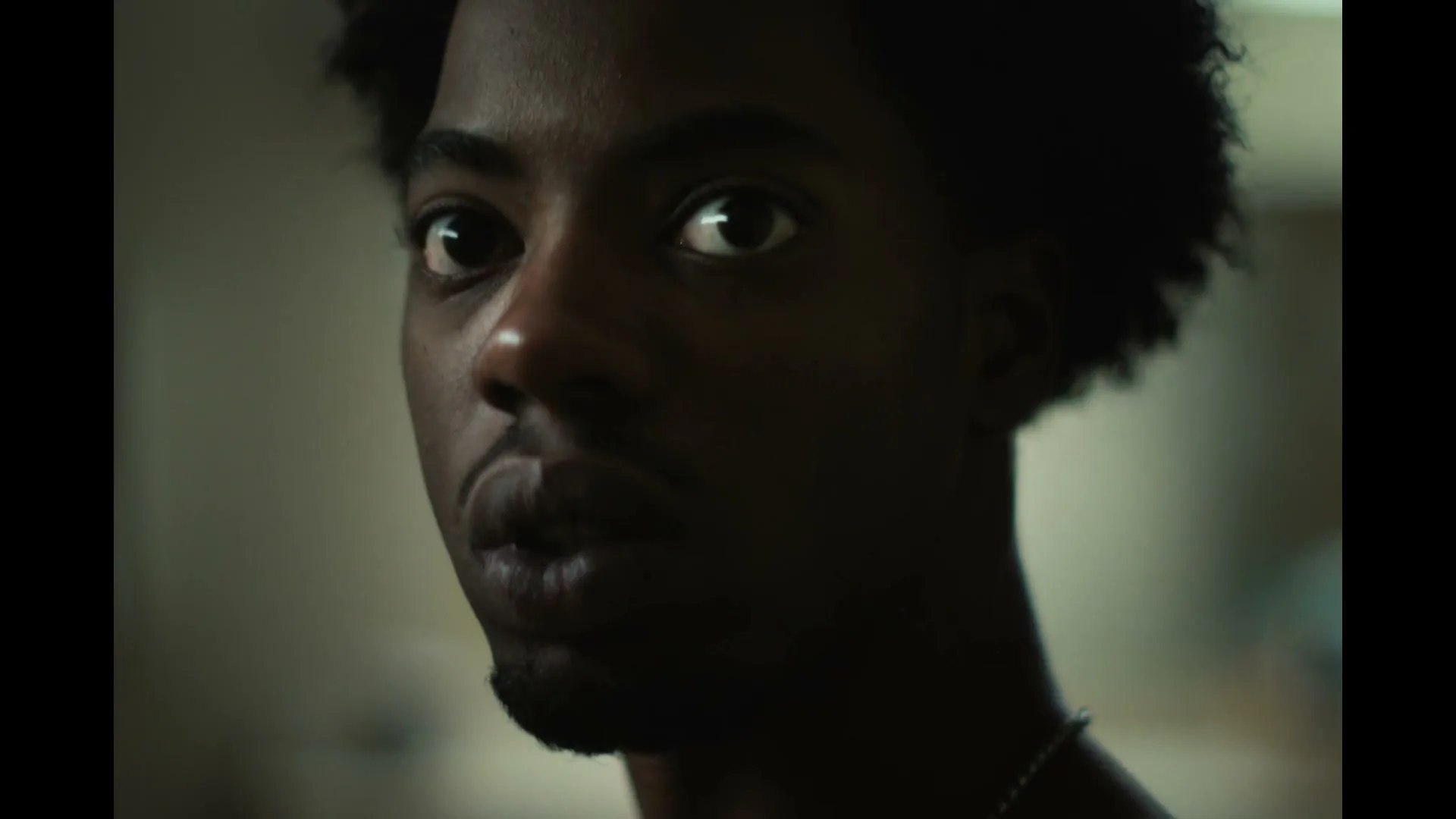 A young Black man with dark curly hair and a goatee looks directly at the camera with wide eyes, appearing startled in a dimly lit room.
