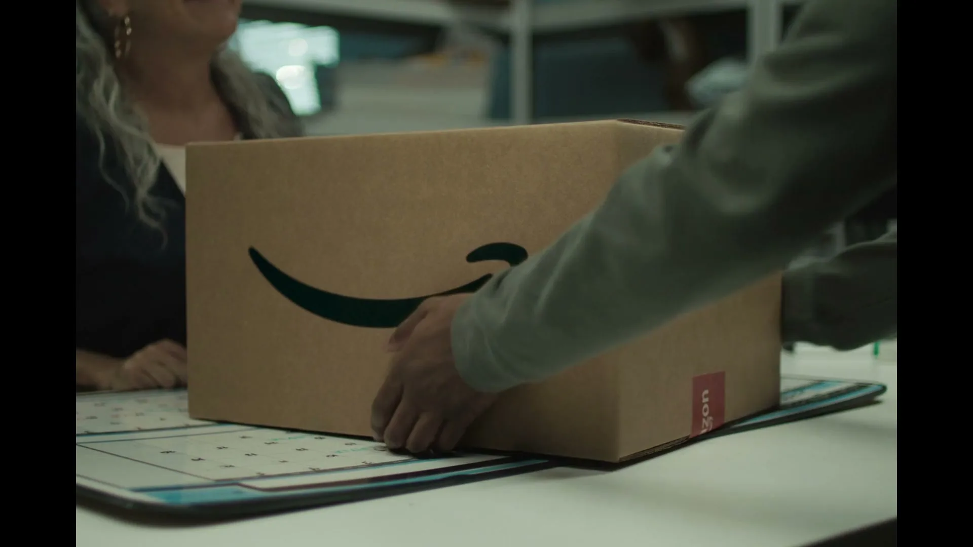 A person's hands pick up an Amazon package from a desk, with another person smiling in the background.