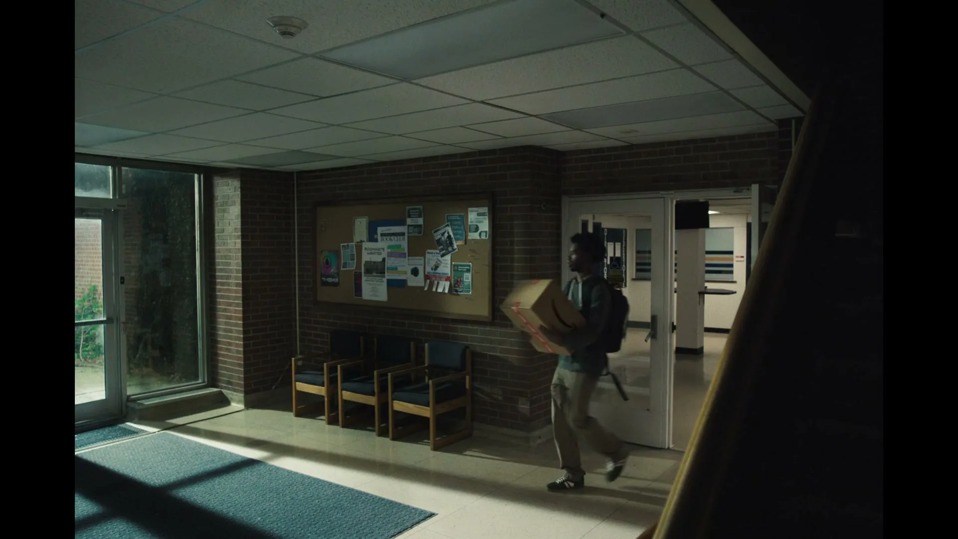 A young man with a backpack carries a large Amazon box through a dimly lit school hallway, walking past a bulletin board and chairs.