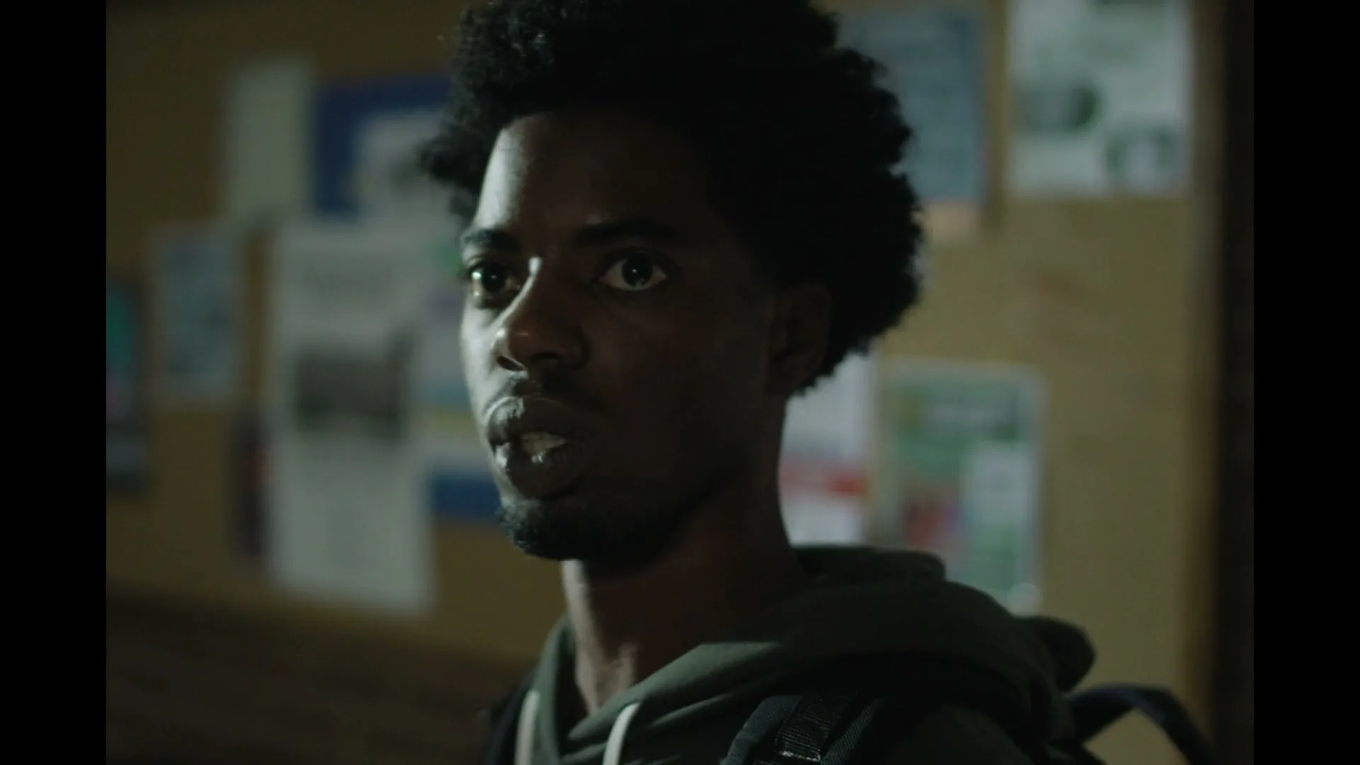 A young Black man with an afro and beard, wearing a dark hoodie, looks around with a surprised expression in a dimly lit room with a bulletin board in the background.