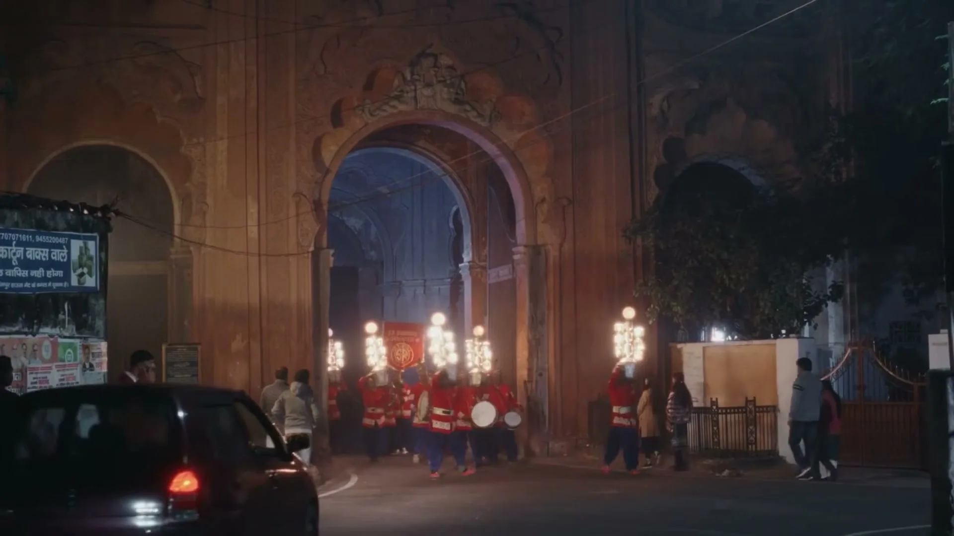 A band of men in red and blue uniforms with drums and bright lights marches out of an ornate archway at night, with a car and other people on the street.