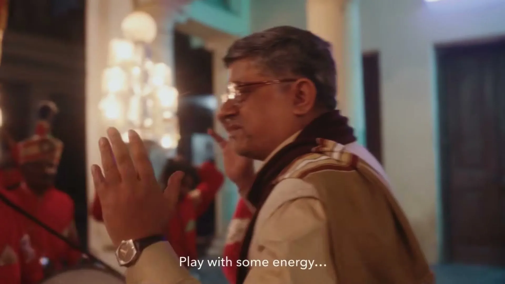 A middle-aged man in glasses and traditional attire conducts a marching band in red uniforms, playing drums and brass instruments at night.