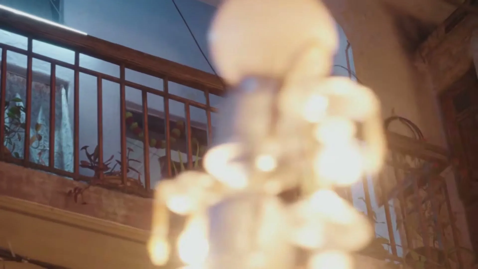 A woman in her 30s holds a baby on a balcony, looking down at something off-screen, with blurred lights in the foreground.