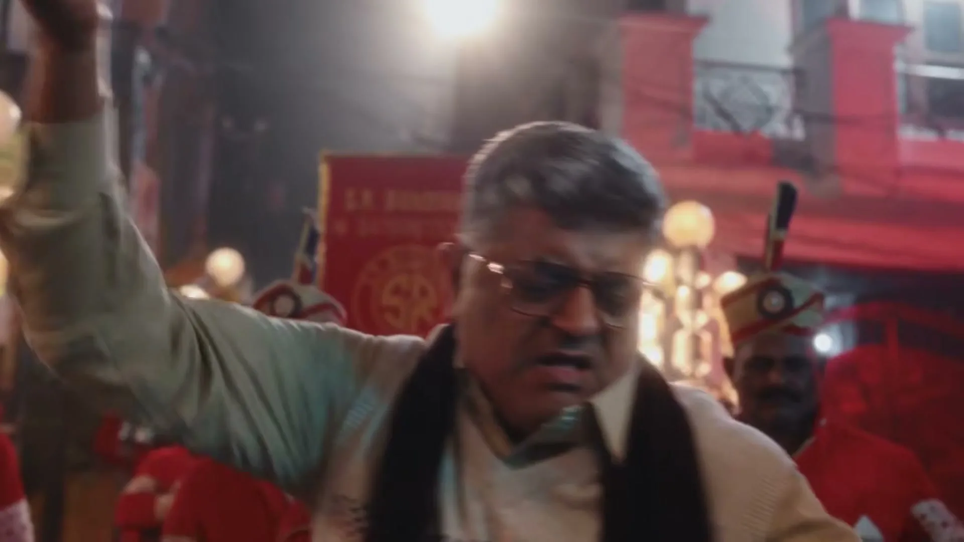 A middle-aged man with glasses and a white shirt dances energetically with his arm raised, surrounded by a band in a festive street at night.