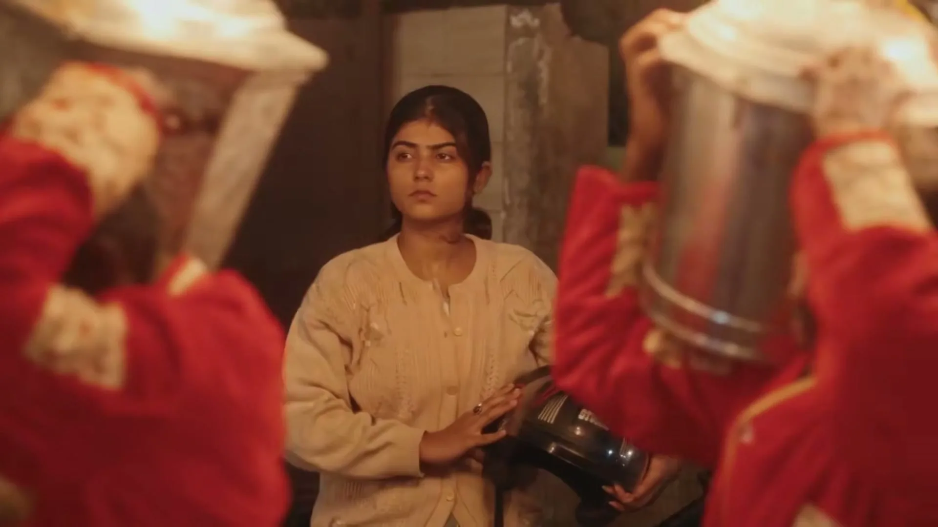 A young woman in a cream sweater stands between two band members in red uniforms, holding a helmet, looking serious at night.